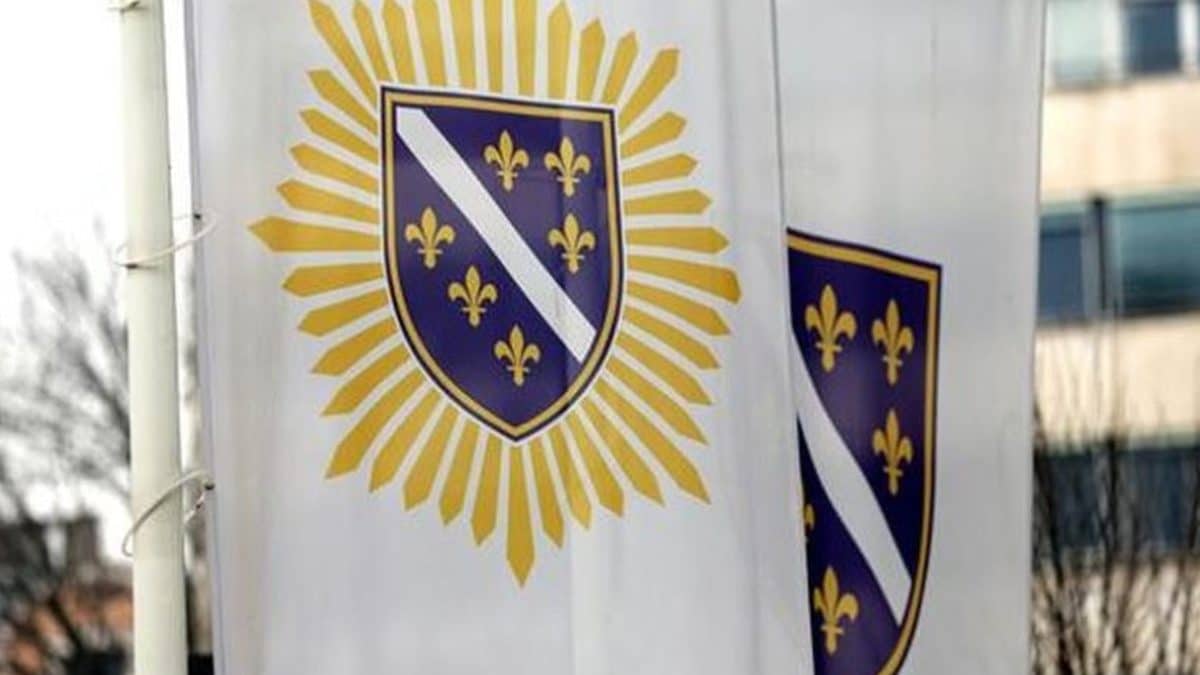 Zastava Armije BiH s ljiljanima i štitom ispred zgrade, simbol bivše vojne strukture u ratu u Bosni i Hercegovini.