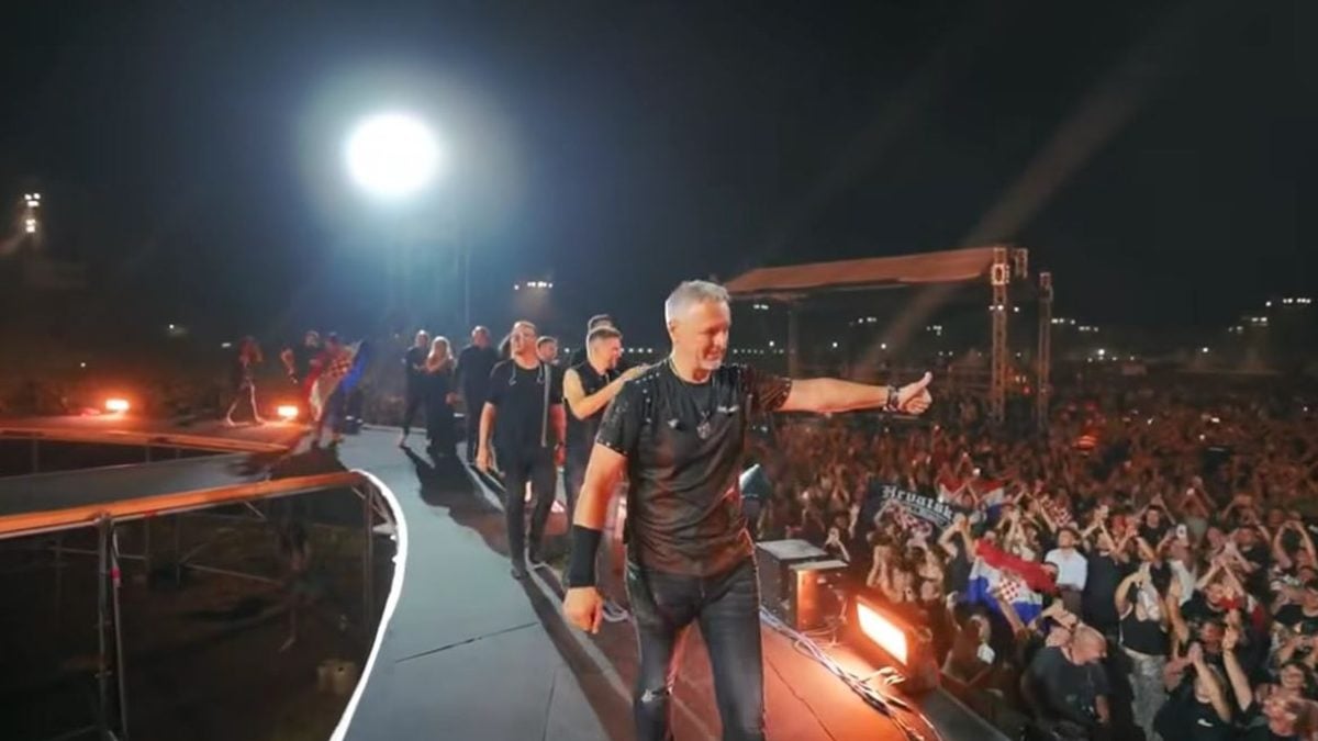 Marko Perković Thompson pozdravlja publiku s pozornice tijekom rekordnog koncerta na Hipodromu u Zagrebu 5. srpnja 2025., dok iza njega stoje članovi benda i tisuće okupljenih obožavatelja s hrvatskim zastavama.