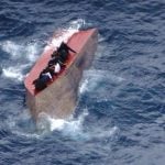 Migranti sjede na prevrnutom brodu uz obalu Libije nakon nesreće u središnjem Sredozemlju.